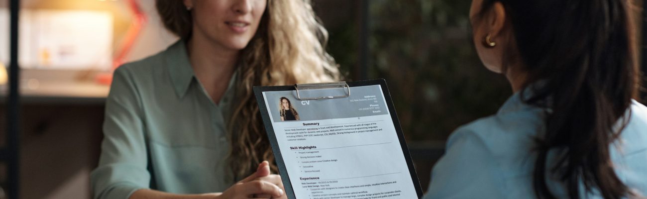 Rear view of businesswoman examining resume of young girl while she sitting at table and telling about herself at interview